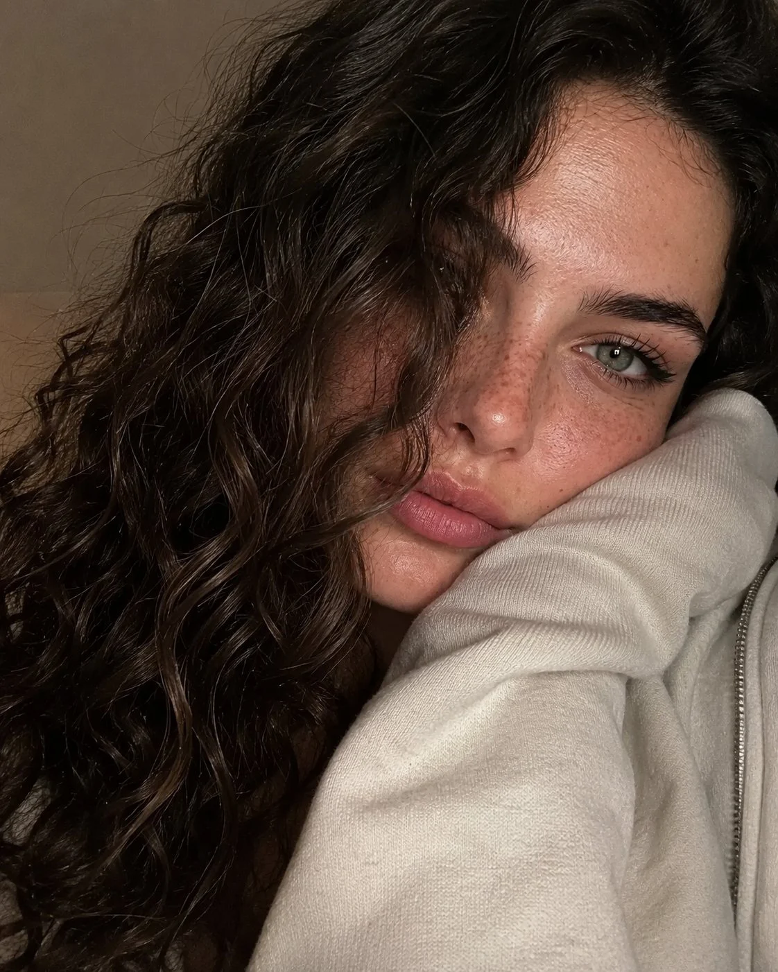 Intimate close-up selfie-style portrait of a woman resting her cheek on the shoulder of a cream hoodie against a warm grey wall.