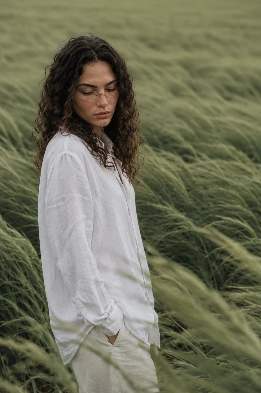 Woman in an oversized white linen shirt standing in a vast field of tall windswept green grass under flat overcast light.