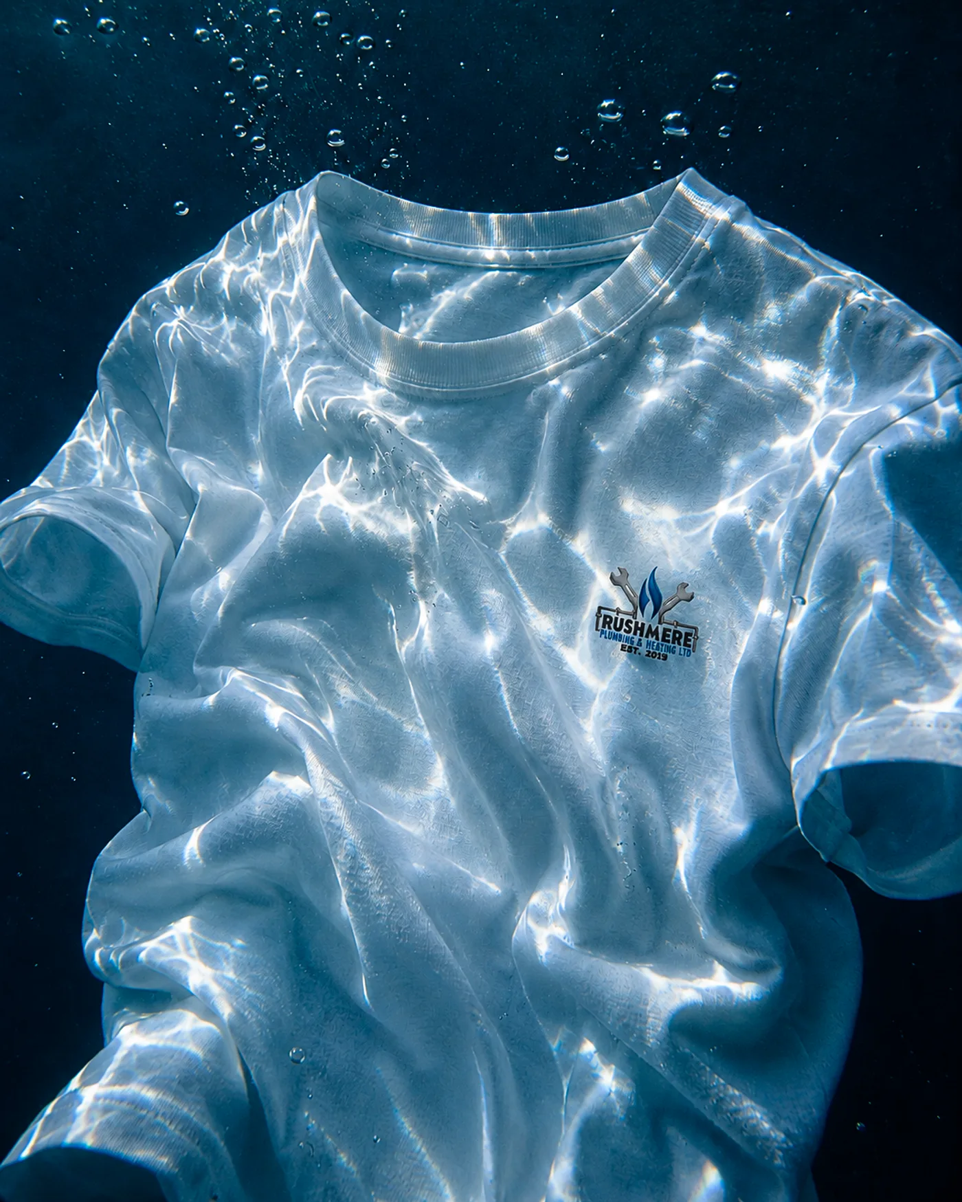 White Rushmere workwear tee suspended underwater, the full colour lockup printed at the chest, caustic light across the fabric